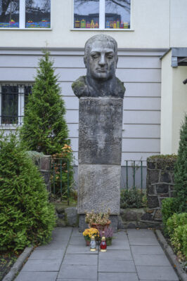 The photo, taken straight on, depicts a bust of a man made of gray stone. It is placed on a tall, rectangular pedestal. The figure has a serious expression and is looking straight ahead. His short-cropped hair does not cover his ears. Beneath the bust, part of the pedestal on which it stands is visible. On the wall of the rectangular block, an inscription is carved in a simple font: “Alf Liczmański. Umęczony 20 marca 1940” (the month is written in Roman numerals). Below the inscription, in the central part of the pedestal, a scout cross is carved in the form of an equilateral cross. On the horizontal arms of the badge, the word “Czuwaj” is visible.