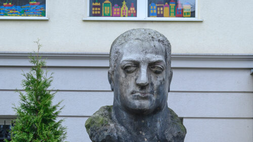 The photo, taken straight on, depicts a bust of a man made of gray stone. It is placed on a tall, rectangular pedestal. The figure has a serious expression and is looking straight ahead. His short-cropped hair does not cover his ears. Beneath the bust, part of the pedestal on which it stands is visible. On the wall of the rectangular block, an inscription is carved in a simple font: “Alf Liczmański. Umęczony 20 marca 1940” (the month is written in Roman numerals). Below the inscription, in the central part of the pedestal, a scout cross is carved in the form of an equilateral cross. On the horizontal arms of the badge, the word “Czuwaj” is visible.