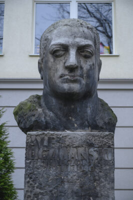 The photo, taken straight on, depicts a bust of a man made of gray stone. It is placed on a tall, rectangular pedestal. The figure has a serious expression and is looking straight ahead. His short-cropped hair does not cover his ears. Beneath the bust, part of the pedestal on which it stands is visible. On the wall of the rectangular block, an inscription is carved in a simple font: “Alf Liczmański. Umęczony 20 marca 1940” (the month is written in Roman numerals). Below the inscription, in the central part of the pedestal, a scout cross is carved in the form of an equilateral cross. On the horizontal arms of the badge, the word “Czuwaj” is visible.