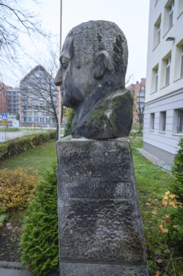 The photo, taken straight on, depicts a bust of a man made of gray stone. It is placed on a tall, rectangular pedestal. The figure has a serious expression and is looking straight ahead. His short-cropped hair does not cover his ears. Beneath the bust, part of the pedestal on which it stands is visible. On the wall of the rectangular block, an inscription is carved in a simple font: “Alf Liczmański. Umęczony 20 marca 1940” (the month is written in Roman numerals). Below the inscription, in the central part of the pedestal, a scout cross is carved in the form of an equilateral cross. On the horizontal arms of the badge, the word “Czuwaj” is visible.
