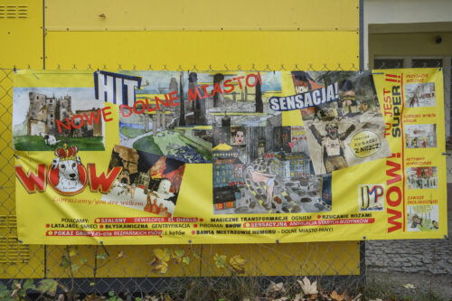 Zdjęcie przedstawiające intensywnie żółty, poziomy banner z nadrukowanym kolażem składający się z różnych rysunków oraz napisów. Do każdego z napisów został zastosowany inny font. Można przeczytać na nim takie zawołania jak: „Nowe Dolne Miasto!”, „Sensacja!”, czy „Tu jest super!!!”. Baner zawieszony jest na zielonej siatce ogrodzeniowej.