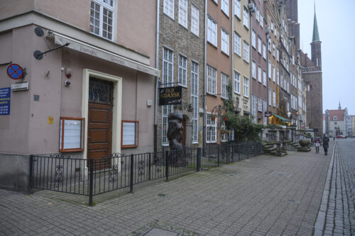 A series of tenement houses at Piwna street in Gdańsk, the first three are captured by a shared, low, metal balustrade; at the second from the left a wooden sculpture of a woman.