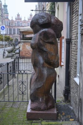 the left side of a wooden sculpture, a woman's body line composed of a double S, a lump of flowing hair at the back of her head; the buildings on Piwna Street in the background.