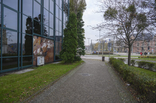 Zdjęcie poziome ukazujące chodnik i trawę, po lewej stronie fragment przeszklonego nowoczesnego budynku, w głębi, na dole budynku widoczna abstrakcyjna mozaika, ułożona z wielobarwnych kafli ceramicznych o róźnorakich kształtach.