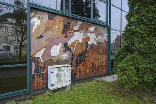 Zdjęcie poziome, z ukosa ukazujące zbliżenie na mozaikę. Kompozycjamozaiki jest diagonalna, abstrakcyjna, złożona z nieregularnych pól koloru, wykonanych z nieregularnych drobnych kafli ceramicznych. Kolorystyka stonowana, brązowa, czarna i różowa, z kontrastującymi białymi plamami.