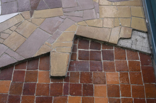 Zdjęcie przedstawia zbliżenie na fragment mozaiki; widoczne dwie warstwy drobnych kafli o nieregularnych kształtach, głębsza warstwa ciemniejsza, w tonacji rudej, na wierzchu jaśniejsze elementy ceramiczne ułożone w kompozycję abstrakcyjną.