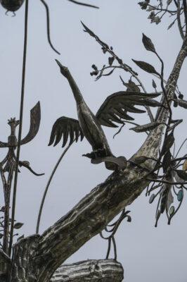 Zdjęcie wykonane z ukosa, od dołu, ze zbliżeniem na jeden z elementów rzeźby w przestrzeni miejskiej. Przypomina on swoim wyglądem kaczkę znajdującą się wśród gałęzi drzewa z pojedynczym liśćmi. Ptak został uchwycony w trakcie przygotowywania się do wzbicia się do lotu. Rzeźba oraz jej elementy zostały wykonane ze stali.