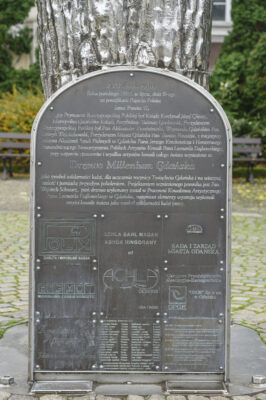 Zdjęcie pionowe przedstawiające ciemnoszarą, prostokątną tablicę o łukowym zwieńczeniu, w ciemnoszarym kolorze. Została ona wykonana z metalu a jej krawędzie są dodatkowo obramowane jasnoszarym wariantem tego materiału. Tablica pokryta jest tekstem objaśniającym oraz logotypami producentów