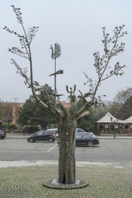 Zdjęcie przedstawia rzeźbę drzewa w przestrzeni miejskiej, wykonaną ze stali. Na jej gałęziach znajdują się pojedyncze liście, zaś pośród nich można dostrzec przedstawicieli różnych gatunków ptactwa, np. kaczkę lub pawia.