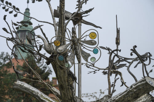 Zdjęcie wykonane z ukosa, od dołu, ze zbliżeniem na jeden z elementów rzeźby w przestrzeni miejskiej. Przypomina on swoim wyglądem dużego motyla znajdującego się wśród gałęzi drzewa z pojedynczym liśćmi. Skrzydła owada zostały ozdobione sześcioma, średniej wielkości emaliami, a na każdej ich parze znajduje się inny kolor dekoracji. Na górnej parze skrzydeł są dwie emalie w kolorze intensywnie żółtym, na środkowej w kolorze cyjanu, zaś na dolnej w kolorze seledynowym.