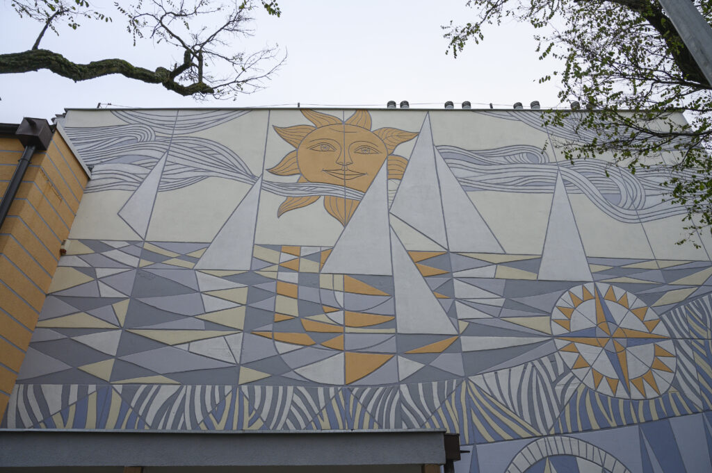 Zdjęcie wykonane od dołu, które przedstawia elewacje ściany budynku, utrzymaną w jasnoszarej i ciemnoszarej tonacji z żółtymi elementami. Swoją kompozycją, na którą stada się wiele figur geometrycznych, nawiązuje ona do sztuki mozaikowej. Wspólnie układają się one w abstrakcyjne przedstawienie sielankowej sceny z białymi żaglówkami na błękitnym jeziorze.
