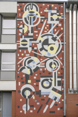 A vertical photo taken in front of it, showing a close-up of one of the walls of the building's façade, the aesthetics of which is reminiscent of mosaic art. There are many geometric figures in bright yellow, blue and gray colors on the red background. In addition, it is also varied with black geometric figures, and the whole brings to mind the works of Wassily Kandinsky from the 1920s
