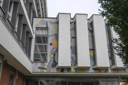Horizontal photo taken from below showing the elevation of the side wall of the projecting entrance to a multi-storey building. Its aesthetics resemble a modernist interpretation of a wall sgraffito. The colors of the façade are kept in bright shades of blue, pink and yellow with gray elements. Its composition has a dynamic and abstract form. The façade composition is separated by five white, longitudinal architectural elements, the side walls of which are intended for window openings.
