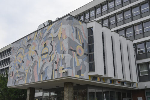 Horizontal photo taken obliquely showing the elevation of the side and front walls of the advanced entrance to the multi-storey building. Its aesthetics resemble a modernist interpretation of a wall sgraffito. The colors of the façade are kept in bright shades of blue, pink and yellow with gray elements. Its composition has a dynamic and abstract form. The composition of the façade on the side of the entrance is separated by five white, longitudinal architectural elements, the side walls of which are intended for window openings.