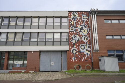 A horizontal photo taken in front of a three-story building, one of the walls with its façade resembling mosaic art. There are many geometric figures in bright yellow, blue and gray colors on the red background. In addition, it is also varied with black geometric figures, and the whole thing brings to mind the works of Wassily Kandinsky from the 1920s.