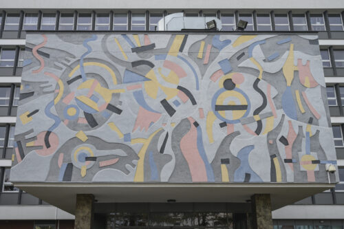 Horizontal photo taken from below showing the frontal wall facade of the advanced entrance to a multi-storey building. Its aesthetics resemble a modernist interpretation of a wall sgraffito. The colors of the façade are kept in bright shades of blue, pink and yellow with gray elements. Its composition has a dynamic and abstract form.