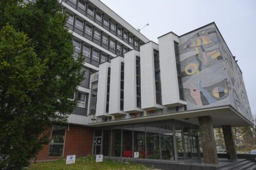 Horizontal photo taken from below showing the elevation of the side wall of the projecting entrance to a multi-storey building. Its aesthetics resemble a modernist interpretation of a wall sgraffito. The colors of the façade are kept in bright shades of blue, pink and yellow with gray elements. Its composition has a dynamic and abstract form. The façade composition is separated by five white, longitudinal architectural elements, the side walls of which are intended for window openings.