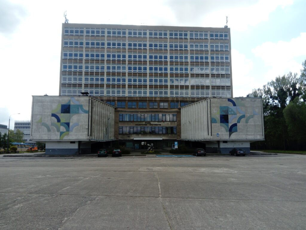 Zdjęcie poziome wykonane na wprost przedstawiające powojenny, wielokondygnacyjny budynek, którego konstrukcja przypomina połączone ze sobą trzy prostopadłościany. Na frontalnych ścianach dwóch z nich znajdują się geometryczne mozaiki utrzymane w niebiesko-błękitno-zielonej kolorystyce.