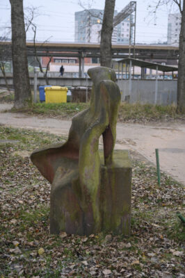 Zdjęcie pionowe wykonane na wprost przedstawiające kamienną rzeźbę, która jest ustawiona na sześciennym postumencie. Figura i postument zostały wykonane z tego samego, ciemnoszarego kamienia a ich powierzchnia zaczyna być porośnięta różnymi porostami. Antropomorficzna figura ma charakter abstrakcyjny i przypomina postać kobiety, która to opiera się na dłoniach o sześcian, głowę zaś ma zarzuconą do tyłu. Granice pomiędzy dwoma bryłami są bardzo zatarte.