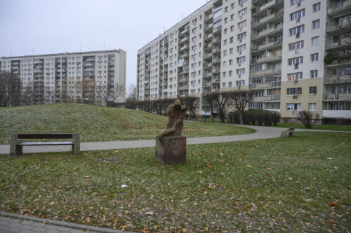 Zdjęcie poziome wykonane na wprost przedstawiające założenie parkowe, otoczone przez architekturę o miejskim charakterze. W centralny punkcie znajduje się kamienna rzeźba, która jest ustawiona na sześciennym postumencie. Figura i postument zostały wykonane z tego samego, ciemnoszarego kamienia a ich powierzchnia zaczyna być porośnięta różnymi porostami. Antropomorficzna figura ma charakter abstrakcyjny i przypomina siedzącą postać kobiety