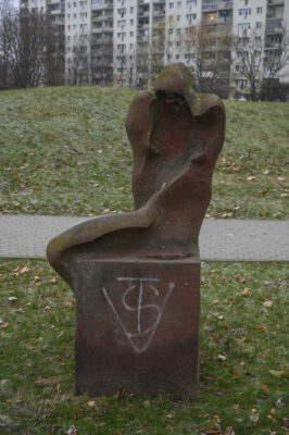 Zdjęcie pionowe wykonane na wprost przedstawiające kamienną rzeźbę ustawioną na sześciennym postumencie. Figura i postument zostały wykonane z tego samego, ciemnoczerwonego kamienia a ich powierzchnia zaczyna być porośnięta różnymi porostami. Antropomorficzna figura ma charakter abstrakcyjny i przypomina zakapturzoną postać. Na ścianie postumentu znajduje się białe, prymitywne graffiti.