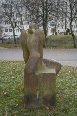 Zdjęcie pionowe wykonane na wprost przedstawiające kamienną rzeźbę ustawioną na sześciennym postumencie. Figura i postument zostały wykonane z tego samego, ciemnoczerwonego kamienia a ich powierzchnia zaczyna być porośnięta różnymi porostami. Antropomorficzna figura ma charakter abstrakcyjny i przypomina zakapturzoną postać, której nakrycie opada aż za ściany postumentu. Granice pomiędzy dwoma bryłami są bardzo zatarte.