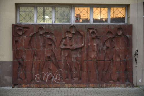 Zdjęcie poziome wykonane na wprost przedstawiające prostokątną instalacje artystyczną, wykonaną z wielu kwadratowych, czerwonych, ceglanych komponentów. Na jej powierzchni znajduje się osiem wklęsłych figur ludzkich ukazanych w bardzo uproszczony, schematyczny sposób – jedynie dostrzegalna jest ich budowa anatomiczna. Cztery postaci męskie i cztery postaci żeńskie zostały przedstawione w momencie wzajemnych interakcji. W lewym dolnym rogu znajduje się białe, prymitywne graffiti. Instalacja stoi na niskim, ceglanym postumencie.