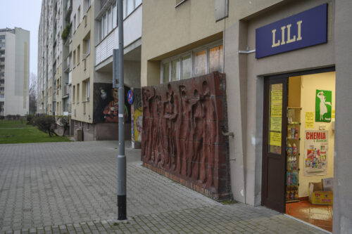 Zdjęcie poziome wykonane z ukosa przedstawiające prostokątną instalacje artystyczną, wykonaną z wielu kwadratowych, czerwonych, ceglanych komponentów. Na jej powierzchni znajduje się osiem wklęsłych figur ludzkich ukazanych w bardzo uproszczony, schematyczny sposób – jedynie dostrzegalna jest ich budowa anatomiczna. Cztery postaci męskie i cztery postaci żeńskie zostały przedstawione w momencie wzajemnych interakcji. W lewym dolnym rogu znajduje się białe, prymitywne graffiti. Instalacja stoi na niskim, ceglanym postumencie.