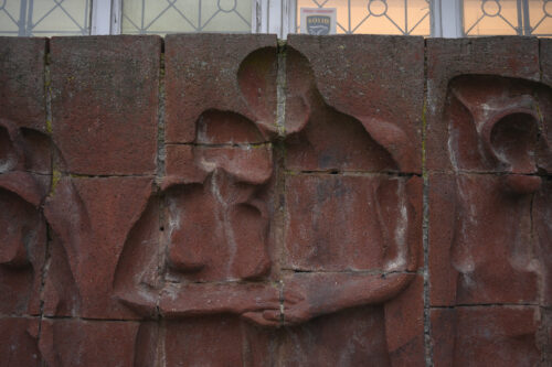 Zdjęcie poziome wykonane na wprost przedstawiające fragment instalacji artystycznej, wykonanej z wielu kwadratowych, czerwonych, ceglanych komponentów. Na jej powierzchni znajdują się wklęsłe figury ludzkie, które zostały ukazane w bardzo uproszczony, schematyczny sposób – jedynie dostrzegalna jest ich budowa anatomiczna. Elementy takie jak piersi czy dłonie podkreślono za pomocą wypukłości. Figury zostały przedstawione w momencie wzajemnych interakcji.