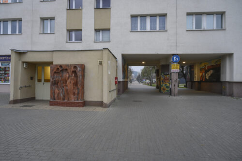 Zdjęcie poziome wykonane z ukosa przedstawiające prostokątną instalacje artystyczną, wykonaną z kilku kwadratowych, czerwonych, ceglanych komponentów. Na jej powierzchni znajdują się trzy, wklęsłe figur ludzkie ukazane w bardzo uproszczony, schematyczny sposób – jedynie dostrzegalna jest ich budowa anatomiczna. Dwie postaci męskie i jedna żeńska zostały przedstawione w momencie wzajemnych interakcji. Instalacja stoi na niskim, ceglanym postumencie i znajduje się obok wejścia do bloku mieszkalnego.