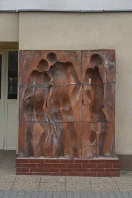 Zdjęcie pionowe wykonane na wprost, przedstawiające prostokątną instalacje artystyczną, wykonaną z kilku kwadratowych, czerwonych, ceglanych komponentów. Na jej powierzchni znajdują się trzy, wklęsłe figur ludzkie ukazane w bardzo uproszczony, schematyczny sposób – jedynie dostrzegalna jest ich budowa anatomiczna. Dwie postaci męskie i jedna żeńska zostały przedstawione w momencie wzajemnych interakcji. Instalacja stoi na niskim, ceglanym postumencie.
