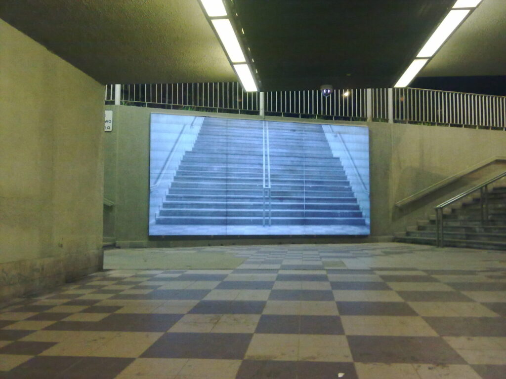 Zdjęcie poziome przedstawiające przejście podziemne, na którego jednej ze ścian jest wyświetlany z rzutnika kadr. Przedstawia on biało-czarne ujęcie prostych schodów z metalową poręczą. Podłoga przejścia została wyłożona ciemnoszarymi i białymi płytami, które wspólnie tworzą wzór szachownicy.