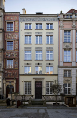 Zdjęcie pionowe, wykonane na wprost, przedstawiające pięciokondygnacyjną kamienicę w tkance zabytkowej Głównego Miasta w Gdańsku. Jej jasnożółta fasada jest ozdobiona prostokątnymi, wielokolorowymi mozaikami a także małymi rzeźbami głów ludzkich.