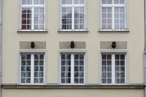 Zdjęcie poziome, wykonane na wprost, ze zbliżeniem na trzy niewielkich rozmiarów rzeźby głów ludzkich, które zdobią jasnożółtą fasadę jednej z kamienic w tkance zabytkowej Głównego Miasta w Gdańsku.