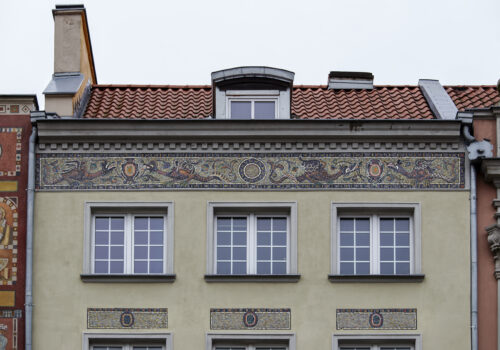 Zdjęcie poziome, wykonane na wprost, ze zbliżeniem na jedną z kilku prostokątnych, wielobarwnych mozaik zdobiących jasnożółtą fasadę jednej z kamienic w tkance zabytkowej Głównego Miasta w Gdańsku.