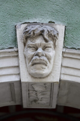 Zdjęcie pionowe ze zbliżeniem na jedną z karykaturalnych płaskorzeźb głów ludzkich znajdujących się w podcieniach arkadowych. Przedstawia ona mężczyznę w średnim wieku z nienaturalnym grymasem twarzy.