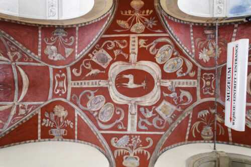 Zdjęcie poziome przedstawiające fresk pokrywający podcienie arkadowe. Została na nim przedstawiona różnorodna flora i fauna w odcieniach beżu i umieszczona na ciemnoczerwonym tle.