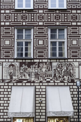 Zdjęcie poziome, wykonane na wprost, ze zbliżeniem dekorację ze sceną historyczną zdobiącą bordowo-brązową fasadę jednej z kamienic w tkance zabytkowej Głównego Miasta w Gdańsku.