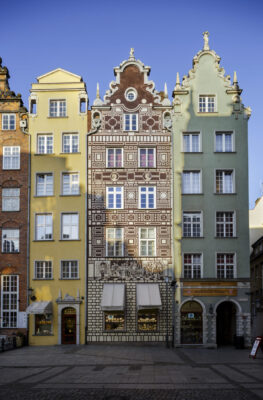 Zdjęcie pionowe, wykonane na wprost, przedstawiające pięciokondygnacyjną kamienicę w tkance zabytkowej Głównego Miasta w Gdańsku. Jej bordowo-brązowa fasada jest ozdobiona graficznym przedstawieniem sceny historycznej a także różnymi popiersiami i pseudoboniowaniem.