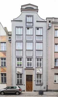 Zdjęcie pionowe, wykonane na wprost, przedstawiające pięciokondygnacyjną kamienicę w tkance zabytkowej Głównego Miasta w Gdańsku. Jej błękitno-szara fasada jest ozdobiona prostą, geometryczną dekoracją, utrzymaną w szarej tonacji.