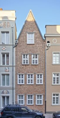 Zdjęcie pionowe, wykonane na wprost, przedstawiające czterokondygnacyjną kamienicę w zabytkowej tkance Głównego Miasta w Gdańsku. Jej ceglana fasada jest ozdobiona delikatnymi, wręcz filigranowymi elementami dekoracyjnymi wykonanymi w technice metaloplastyki.