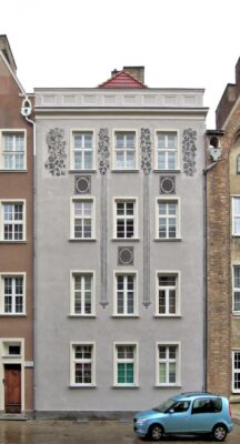 Zdjęcie pionowe, wykonane na wprost, przedstawiające czterokondygnacyjną kamienicę w zabytkowej tkance Głównego Miasta w Gdańsku. Jej szara fasada jest ozdobiona przedstawieniami bujnych akantów a także elementami geometrycznymi.