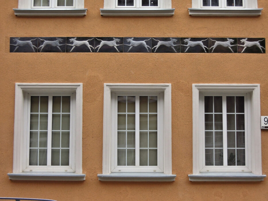 Zdjęcie pionowe, wykonane na wprost, ze zbliżeniem na fragment dekoracji architektonicznej znajdujący się na fasadzie jednej z kamienic w zabytkowej tkance Głównego Miasta w Gdańsku. Pomarańczowa fasada kamienicy została pokryta ceramicznymi płytami z przedstawieniami biegnących psów łownych.