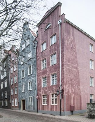 Zdjęcie pionowe, wykonane z ukosa, przedstawiające pięciokondygnacyjne kamienice w zabytkowej tkance Głównego Miasta w Gdańsku. Ciemnoróżowa oraz jasnoniebieska fasada zostały pokryte bardzo rozbudowaną dekoracją graficzną z przedstawieniami ludzkimi, jak również zwierzęcymi i floralnymi. Całość kompozycji sprawia wrażenie chaotycznej i postapokaliptycznej.