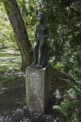 Zdjęcie pionowe, wykonane z ukosa przedstawiające siedzącą figurę ludzką , która została wykonana w brązie. Rzeźba przedstawia całopostaciową figurę kobiety, której anatomiczna budowa ciała została ukazana w sposób naturalny i delikatny. Rzeźba została umieszczona na wysokim, kamiennym, prostopadłościennym postumencie, na którego jednej ze ścian umieszczono plakietkę z napisem: „Przedwiośnie, Anna Pietrowiec”.