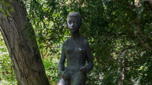 Zdjęcie wykonane z ukosa przedstawiające siedzącą figurę ludzką , która została wykonana w brązie. Rzeźba przedstawia całopostaciową figurę kobiety, której anatomiczna budowa ciała została ukazana w sposób naturalny i delikatny. Rzeźba została umieszczona na wysokim, kamiennym, prostopadłościennym postumencie, na którego jednej ze ścian umieszczono plakietkę z napisem: „Przedwiośnie, Anna Pietrowiec”.