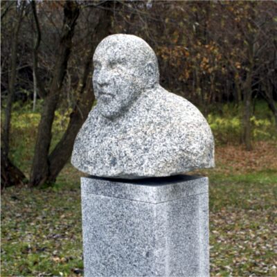 Zdjęcie przedstawiające kamienne popiersi mężczyzny w przestrzeni parkowej. Rzeźba została wykonana z szarego granitu, podobnie jak jej prostopadłościenny postument. Figura przestawia łysego mężczyznę. Jego tors został potraktowany monolityczne, natomiast takie elementy anatomiczne jak: oczy, usta, nos, czy uszy zostały wyraźnie zaznaczone jednak nie są one w pełni ukazane naturalistycznie.