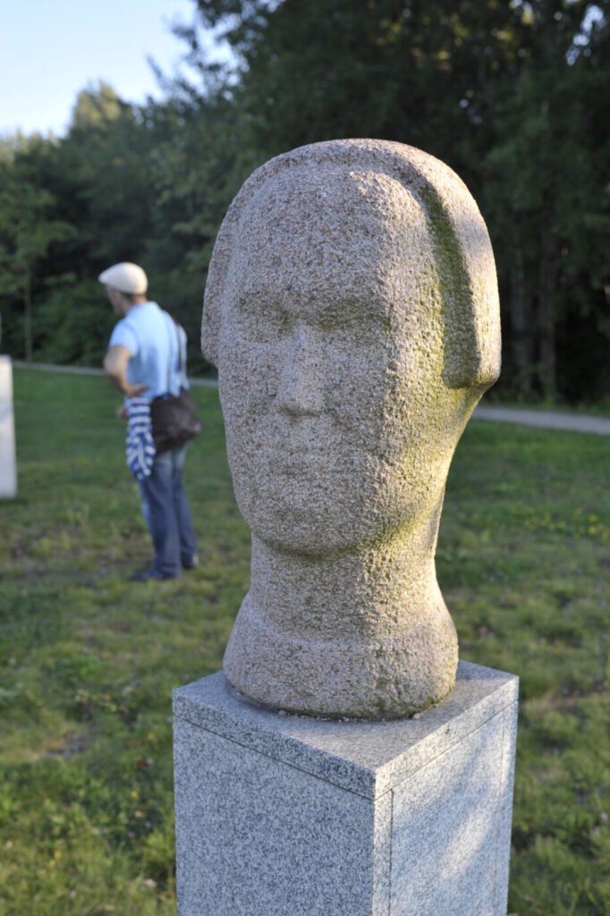 Zdjęcie pionowe przedstawiające kamienna rzeźbę w przestrzeni parkowej. Przedstawia ona głowę kobiety wykonaną w jasnoróżowym kamieniu. Jej rysy twarzy a także elementy anatomiczne takie jak: oczy, nos czy usta zostały zaznaczone pojedynczymi, delikatnymi rytami. Rzeźba ustawiona jest na prostopadłościennym postumencie, który został wykonany z jasnoszarego granitu.