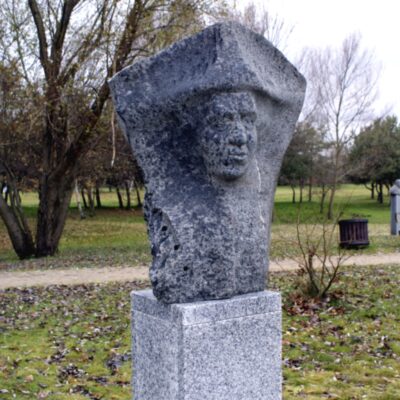 Zdjęcie przedstawiające kamienną, abstrakcyjną rzeźbę w przestrzeni parkowej. Składa się na nią jednorodna, monolityczna bryła, z której wyłania się fragment twarzy przedstawionej postaci. Dostrzegalna jest jedynie jej twarz, a elementy anatomiczne takie jak: nos, usta, podbródek i oczy zostały ukazane w bardzo minimalistyczny sposób. Rzeźba została wykonana w ciemnoszarym kamieniu i ustawiona jest na jasnoszarym, granitowym, prostopadłościennym postumencie.