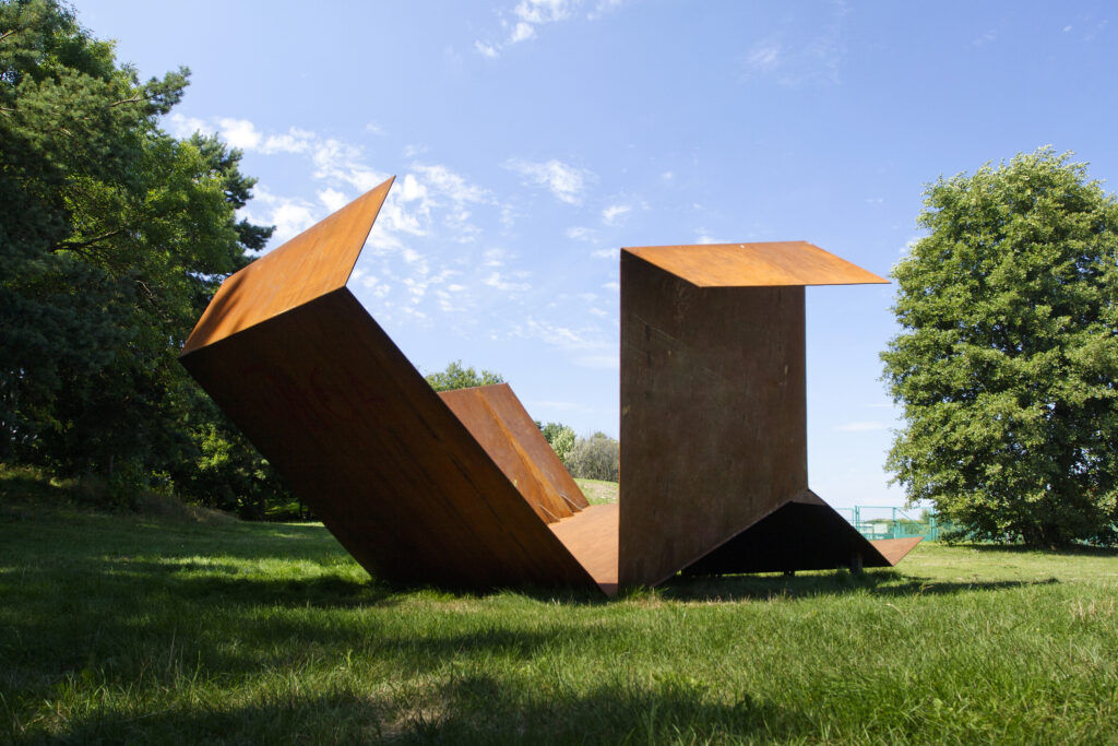 Zdjęcie poziome przedstawiające geometryczną, abstrakcyjną rzeźbę w przestrzeni parkowej. Na jej konstrukcje składa się kilka metalowych prostokątów, które zostały ze sobą wzajemnie połączone pod różnymi kątami. Na ich powierzchniach występuje zaawansowany proces korozji. Cała konstrukcja rzeźby przypomina swoim wyglądem rozłożone pudełko.